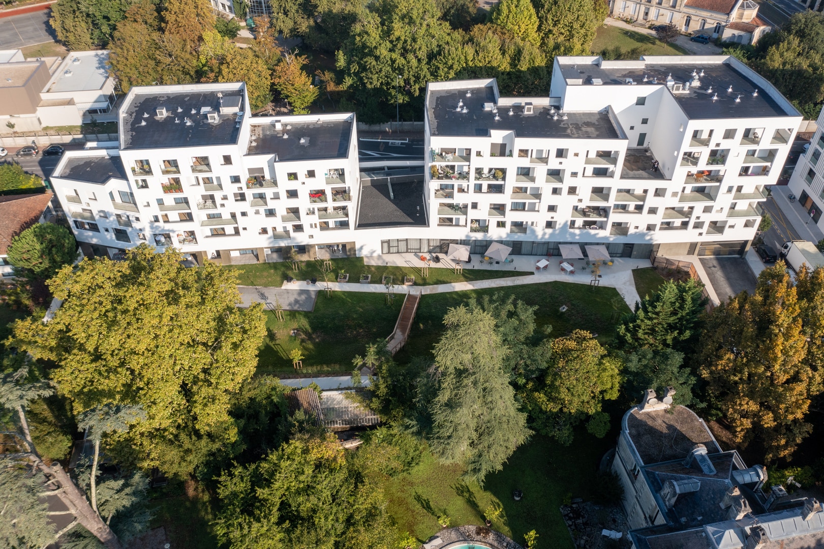 Vue aérienne d'un complexe résidentiel moderne avec un jardin central soigneusement aménagé. Les bâtiments blancs aux fenêtres géométriques contrastent avec le vert luxuriant des arbres environnants, mettant en évidence une architecture urbaine intégrée dans un espace naturel.