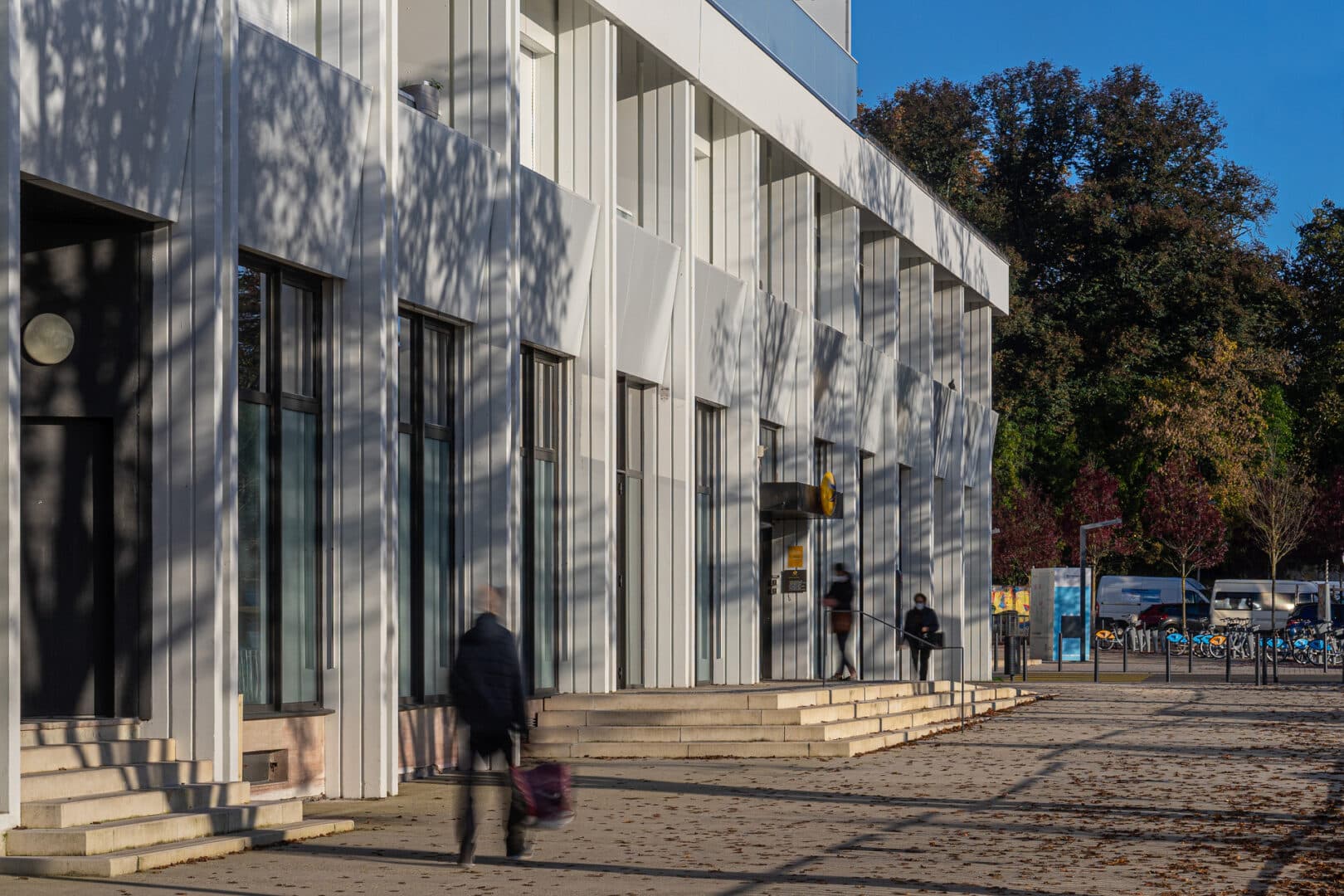Vue extérieure du bâtiment moderne Le Cosmopolitain avec des façades en panneaux blancs et des fenêtres verticales. Des ombres d'arbres se projettent sur le bâtiment par une journée ensoleillée, tandis qu'une personne marche le long du trottoir avec un sac.