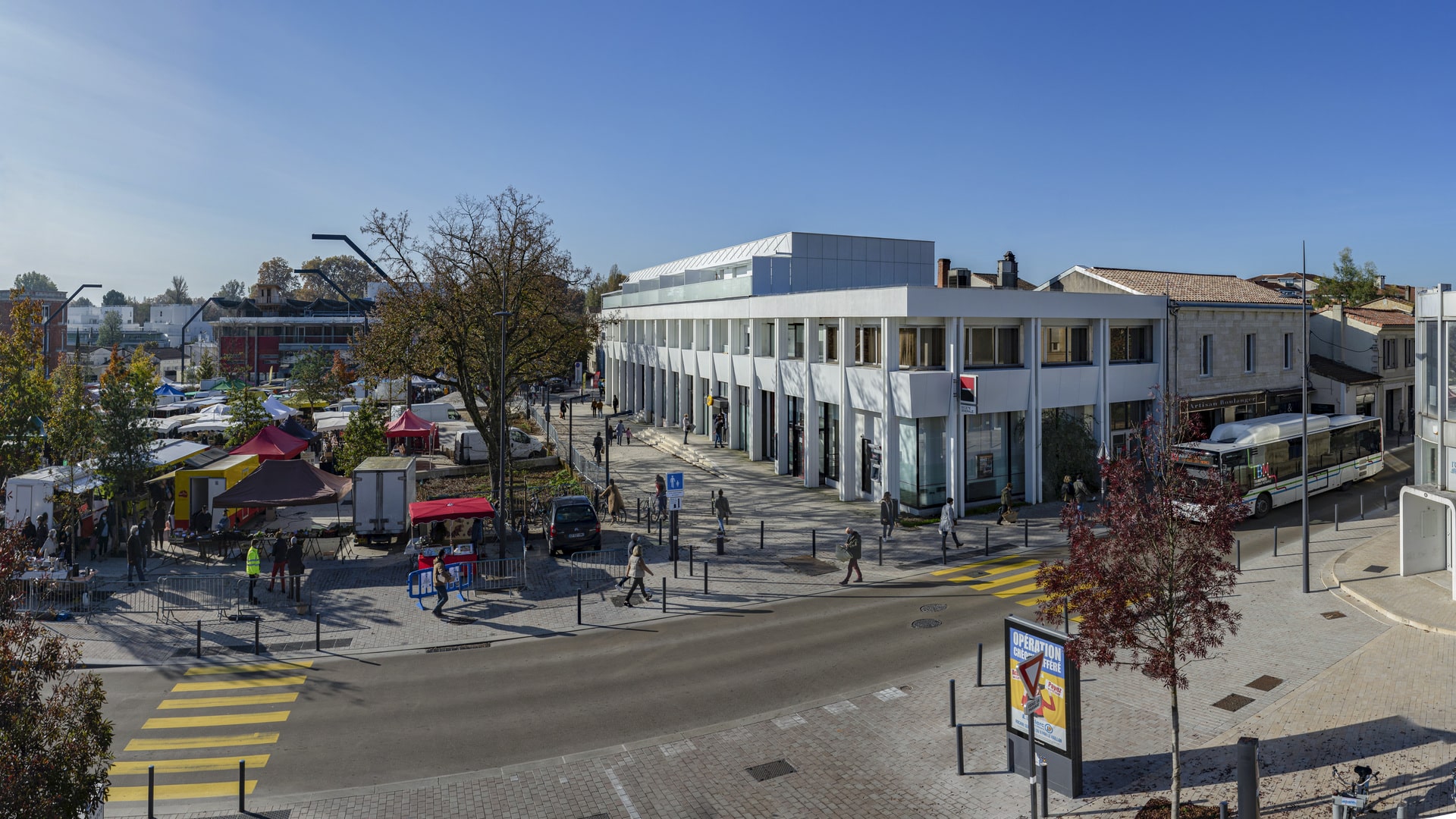 Panorama du quartier urbain avec Le Cosmopolitain à gauche, montrant son architecture contemporaine blanche et sa conception angulaire. Des passants traversent la rue sur un passage piéton, et un marché de plein air occupe l'espace à droite.